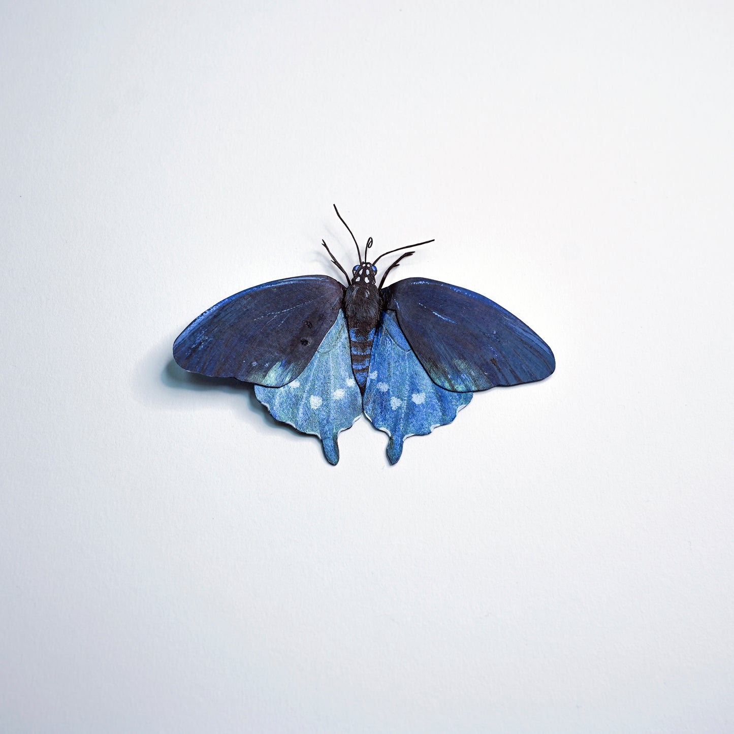 A butterfly-shaped decorative object with dark blue upper wings and lighter blue lower wings with small white spots, centered on a plain light background.