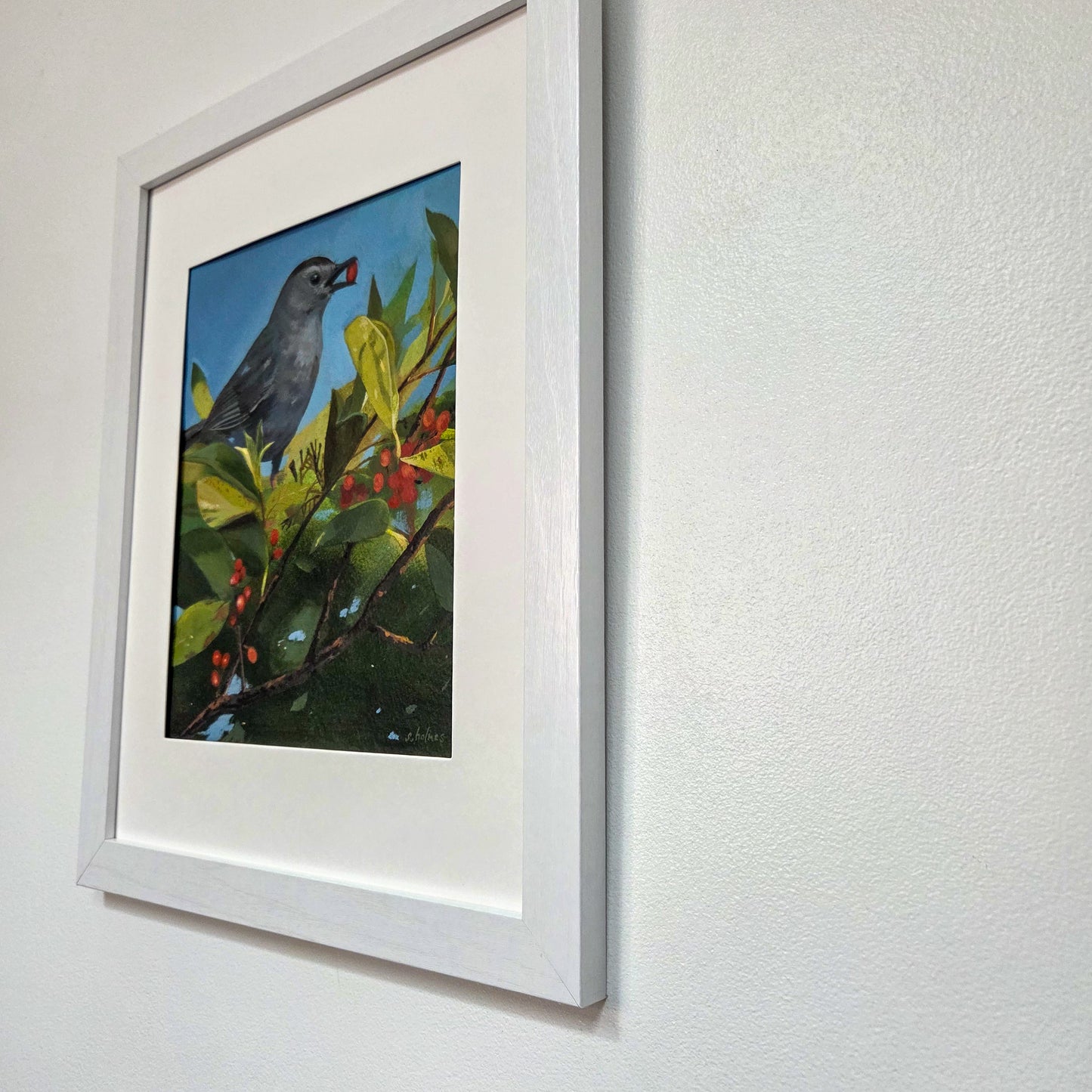 A framed painting of a gray catbird perched on a leafy branch with red berries, displayed on a textured white wall.