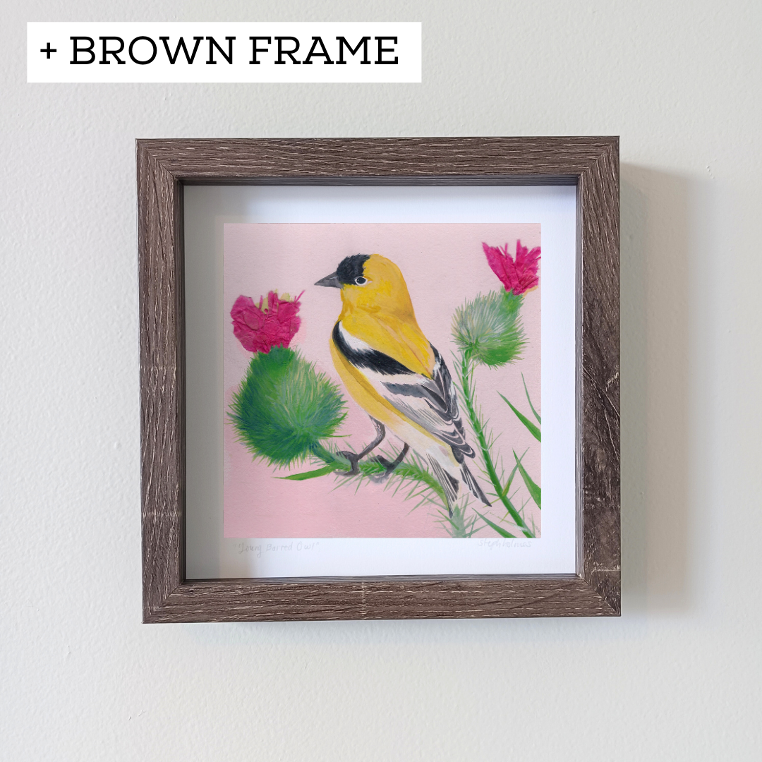 An art print of an American Goldfinch in a brown frame on a white wall.