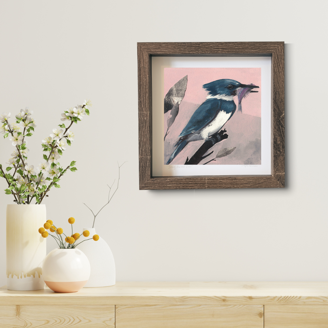 An art print featuring a blue Belted Kingfisher bird in brown frame hanging above a modern wood desk.