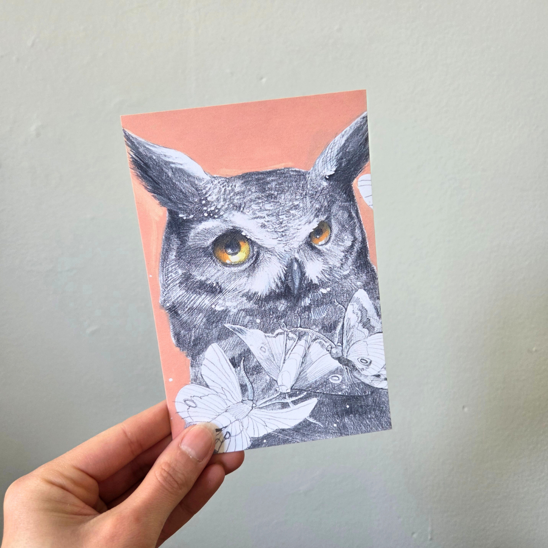 A hand holding an art print postcard featuring a great horned owl.