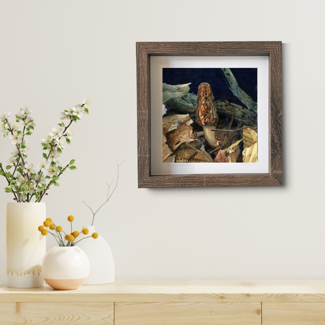 An art print featuring a black morel mushroom in a brown frame on a white wall above a modern wooden desk.