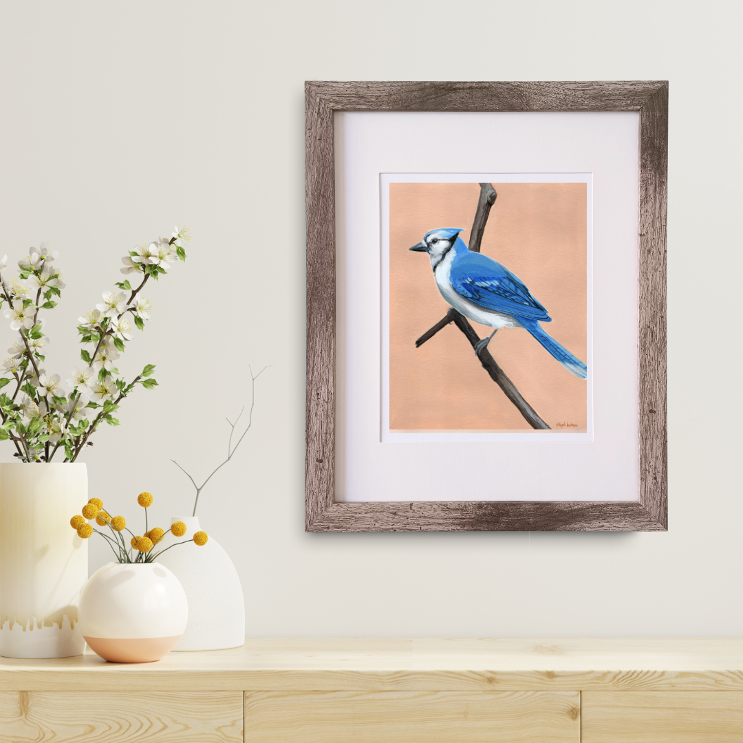 An art print featuring a bright Blue Jay bird in a brown frame hanging above a modern wood table.