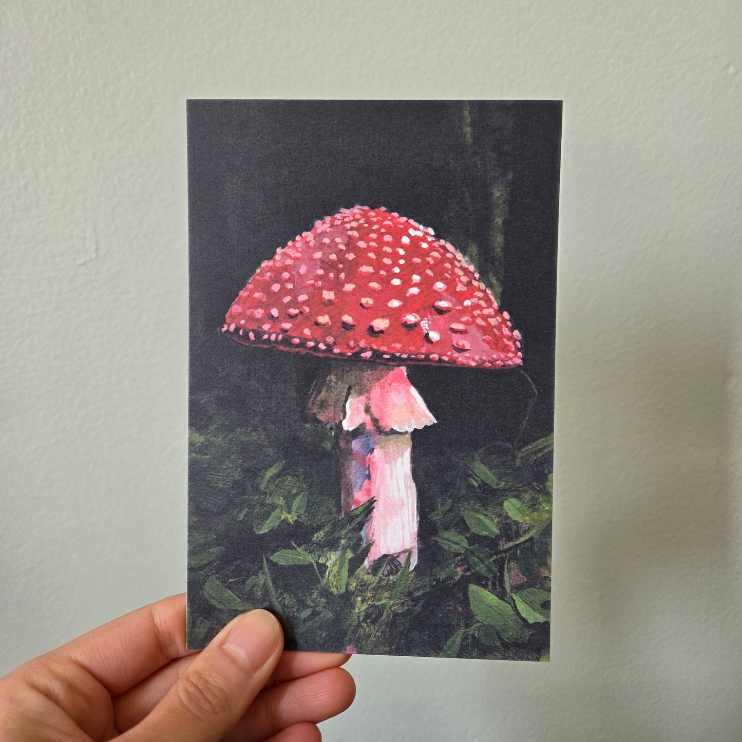 A hand holding an art print postcard featuring a fly amanita red mushroom.