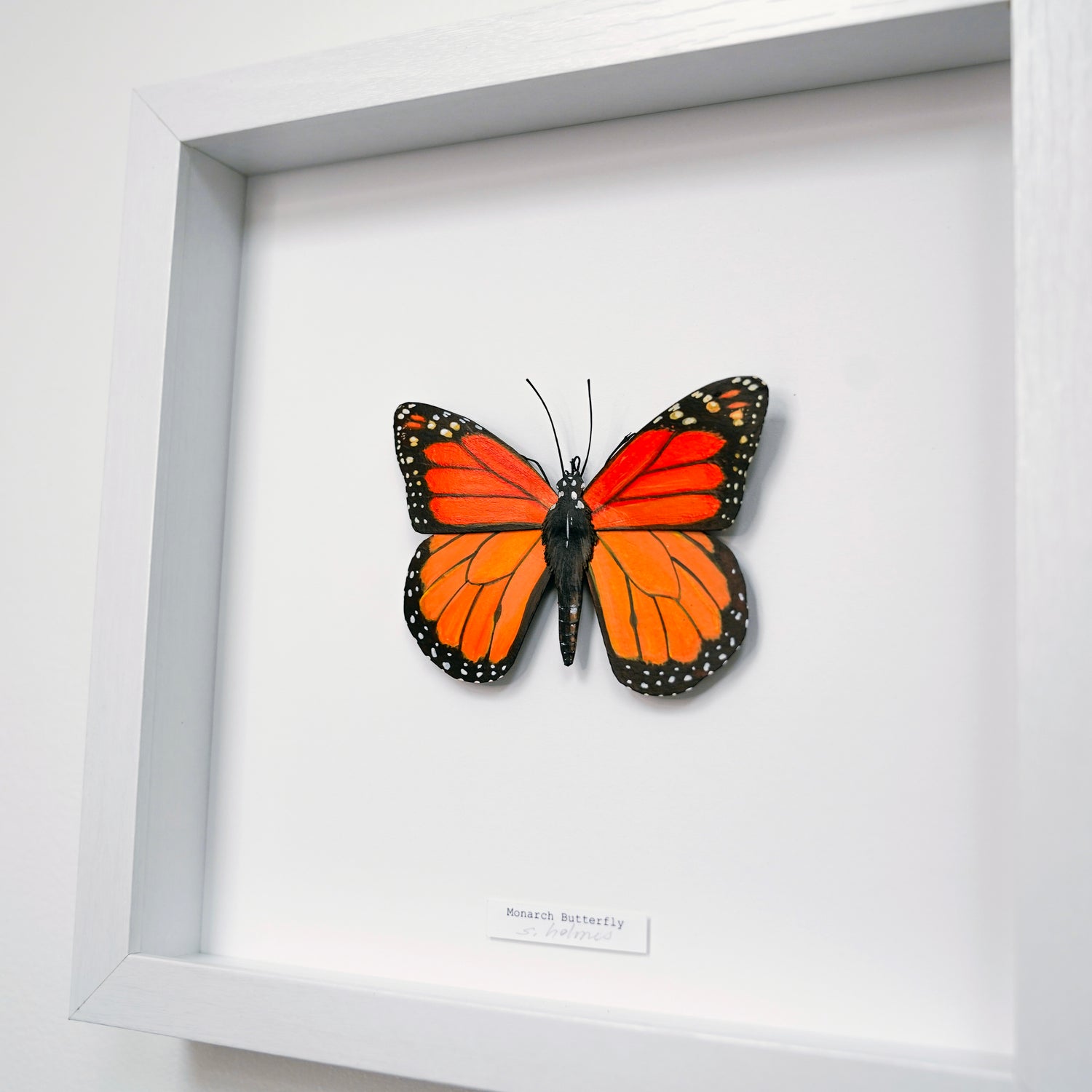 A framed butterfly wall art piece featuring a mounted monarch butterfly with bright orange, red, black, and white patterned wings inside a white shadow box frame.