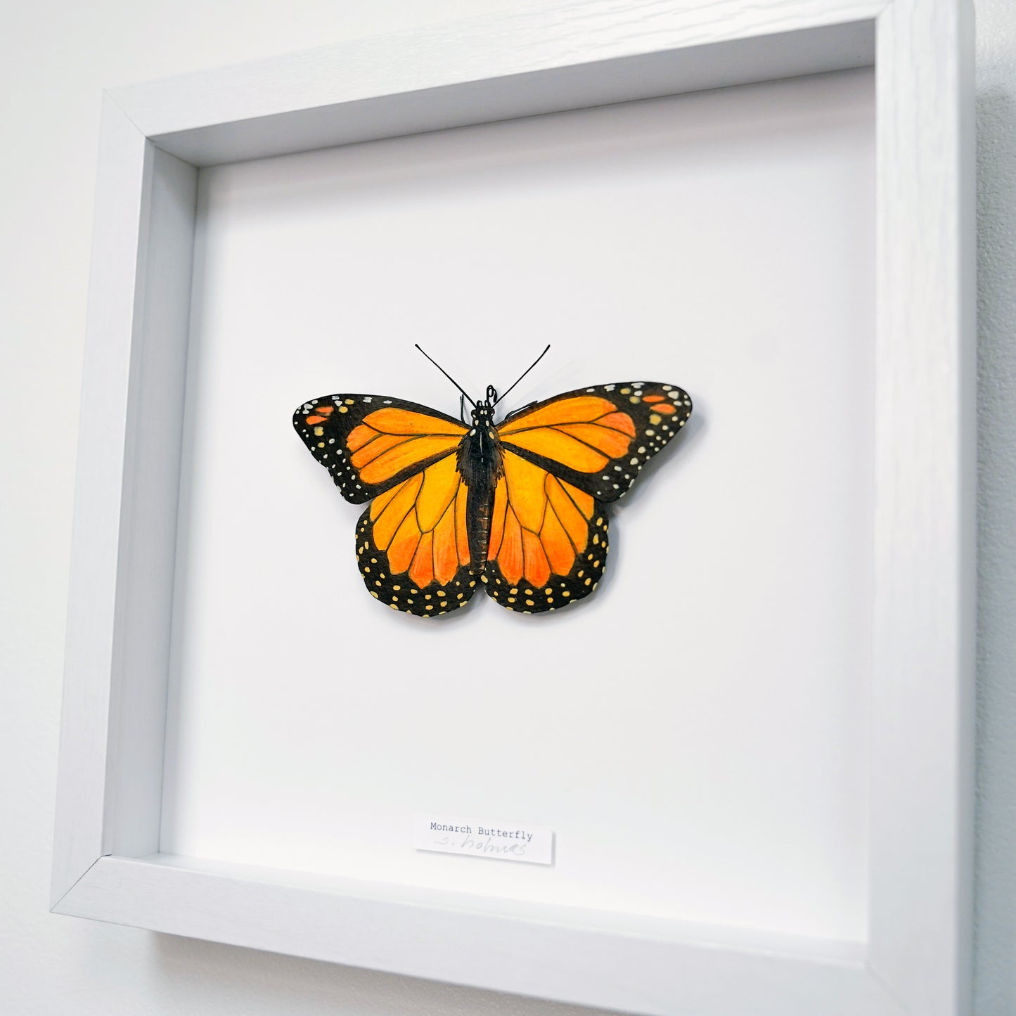 A framed mounted monarch butterfly sculpture on a white background, displayed in a white shadow box frame with a small label at the bottom.