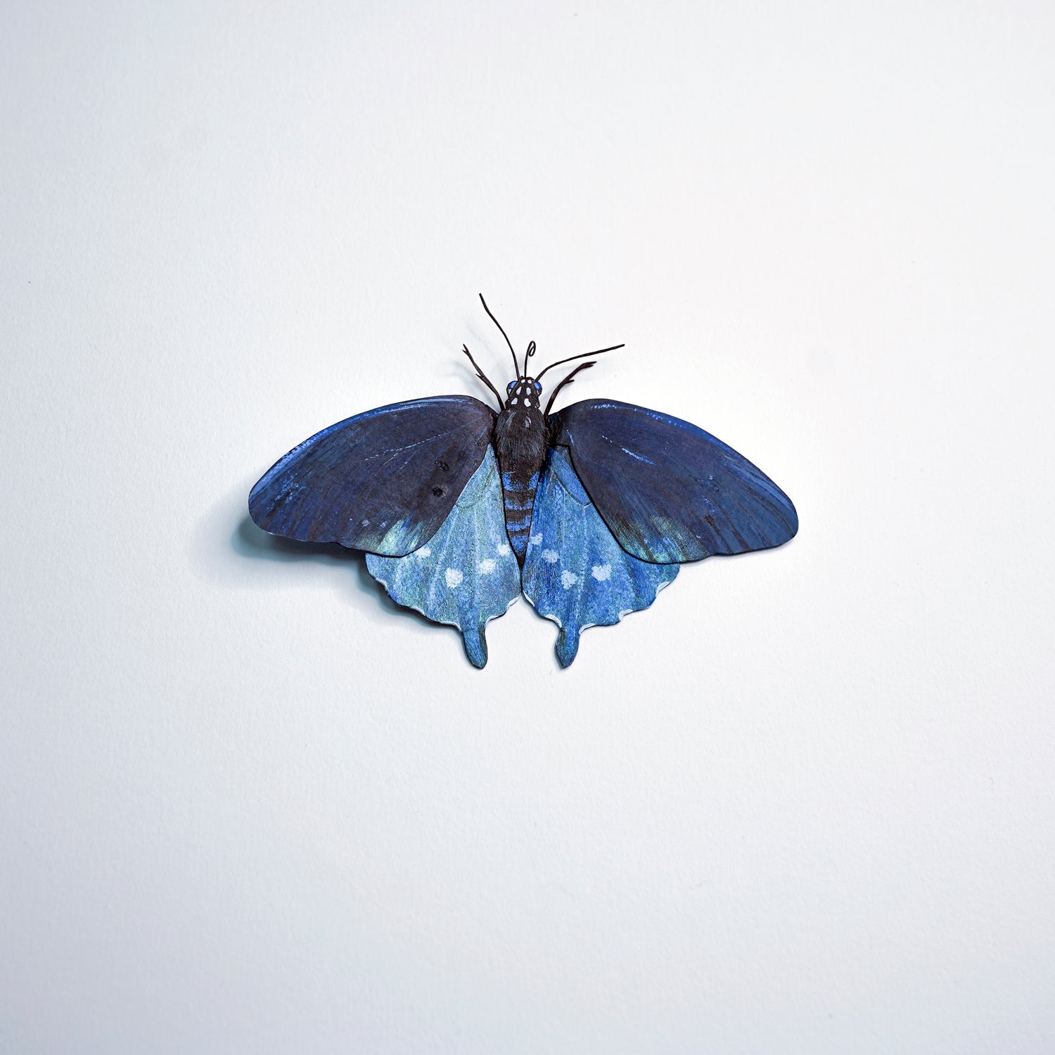 A butterfly-shaped decorative object with dark blue upper wings and lighter blue lower wings with small white spots, centered on a plain light background.