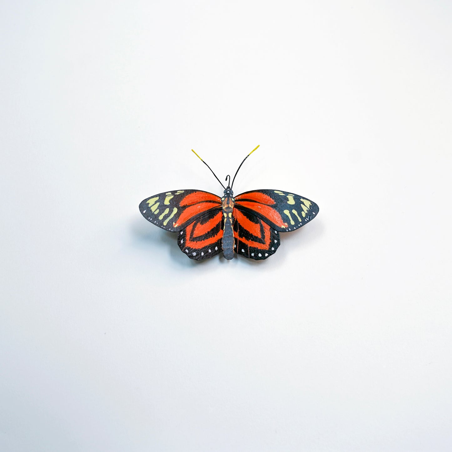 A single butterfly art sculpture centered on a plain white background. The wings are predominantly bright orange-red with black edging and small pale yellow-green accents, plus a dark body and thin yellow antennae.