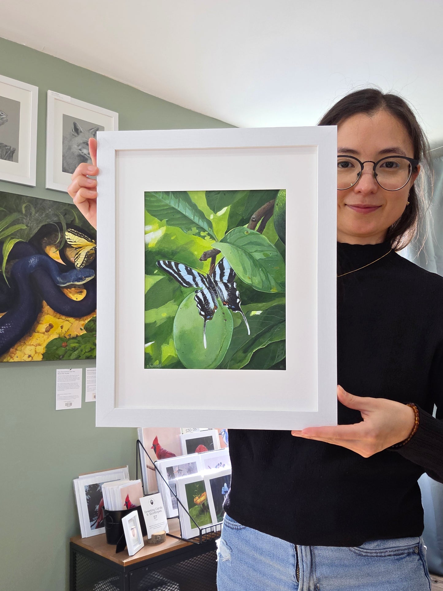 Person holding a framed painting of a butterfly on a leaf with another painting in the background.