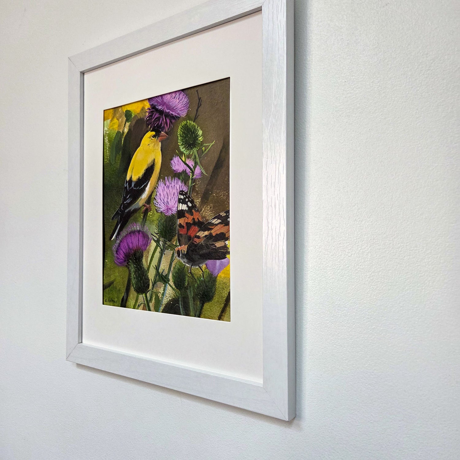A framed art painting featuring a yellow bird perched among purple flowers and green foliage, mounted on a white wall with a wide white mat and light wood frame.