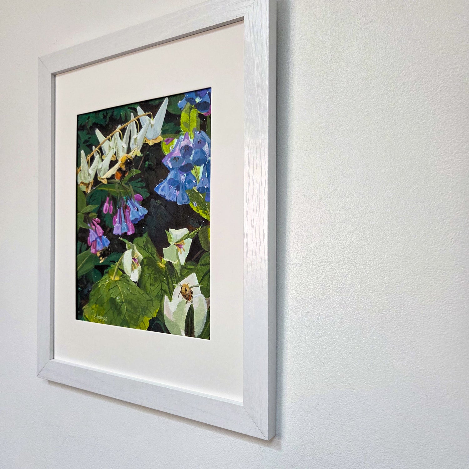 Framed floral art print hanging on a white textured wall. The artwork shows white, blue, and purple flowers with green leaves on a dark background, surrounded by a wide white mat and a white wood frame.