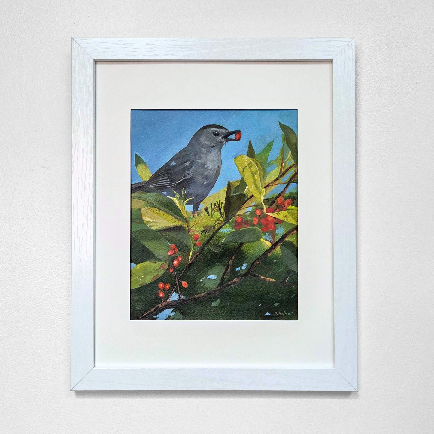 A framed painting featuring a painted gray catbird perched on a branch with green leaves and red berries against a blue sky, set in a white frame on a light wall.