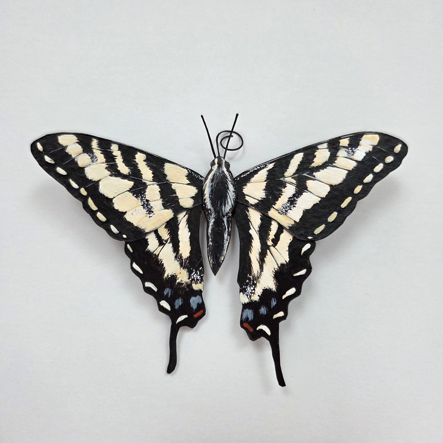 A butterfly art sculpture, an Eastern Tiger Swallowtail, mounted flat against a plain light background. The wings are spread wide, showing a black body and upper wings with bold cream stripes and small red and blue accents near the lower edges.