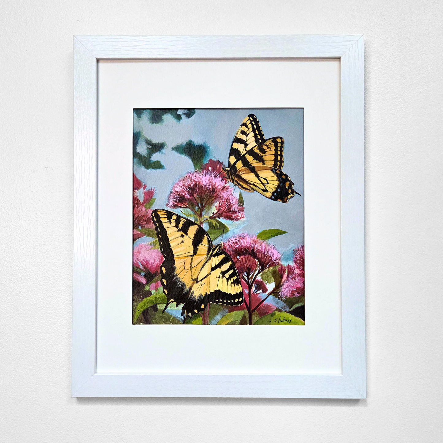 A framed painting featuring two yellow and black swallowtail butterflies perched among pink flowers against a light blue sky, presented in a white frame with a white mat on a light gray wall.