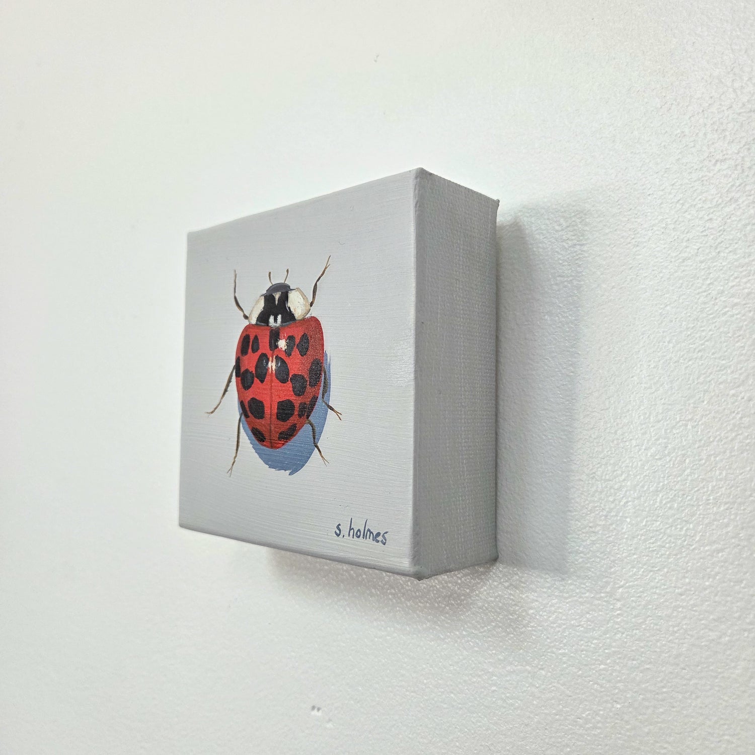 A small square canvas painting of a red ladybug with black spots on a light gray background, mounted on a white textured wall.
