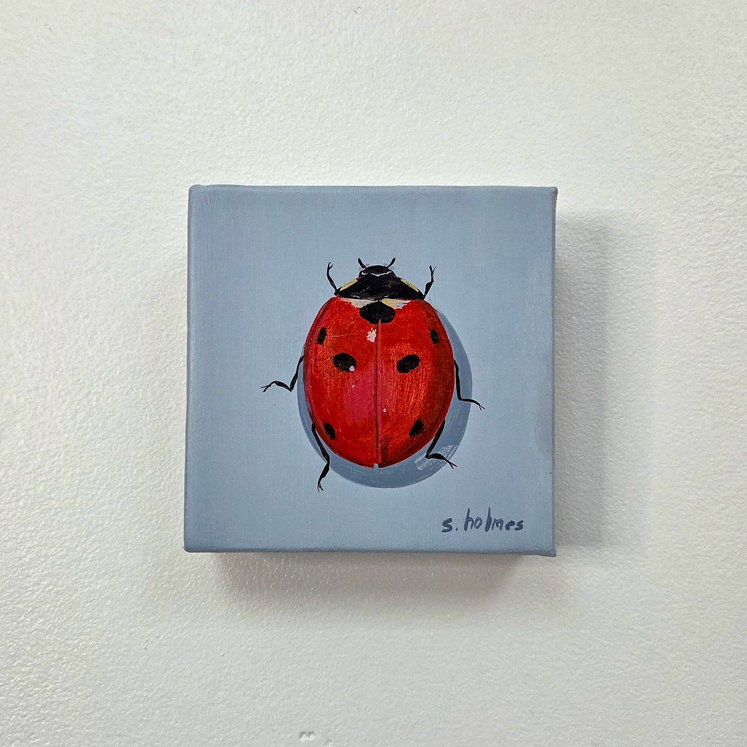 A square canvas painting of a red ladybug with black spots on a light blue-gray background, mounted on a textured off-white wall.