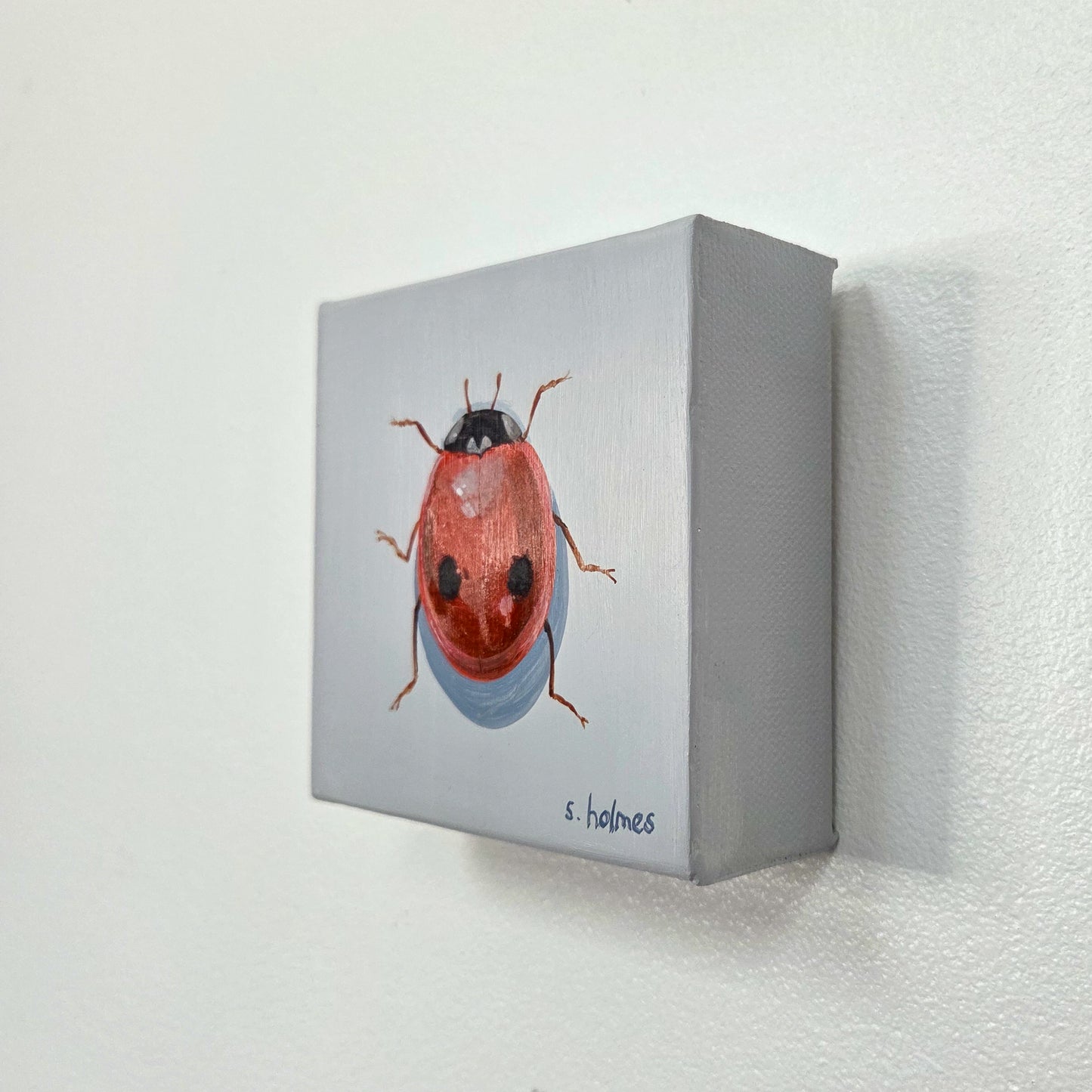 A small square canvas painting of a ladybug mounted on a white textured wall. The canvas has a light gray background with a detailed red, orange, and black ladybug and a blue shadow.
