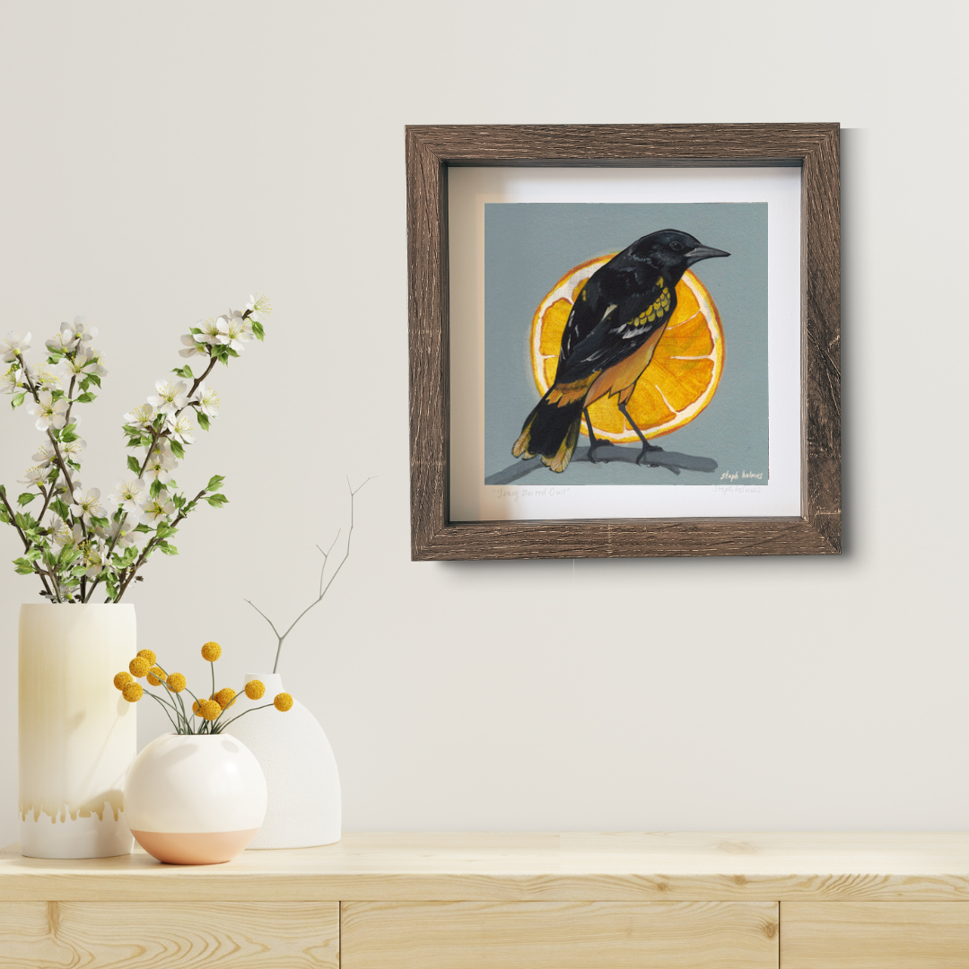 An art print featuring a Baltimore Oriole bird in a brown frame hanging above a modern wood desk.