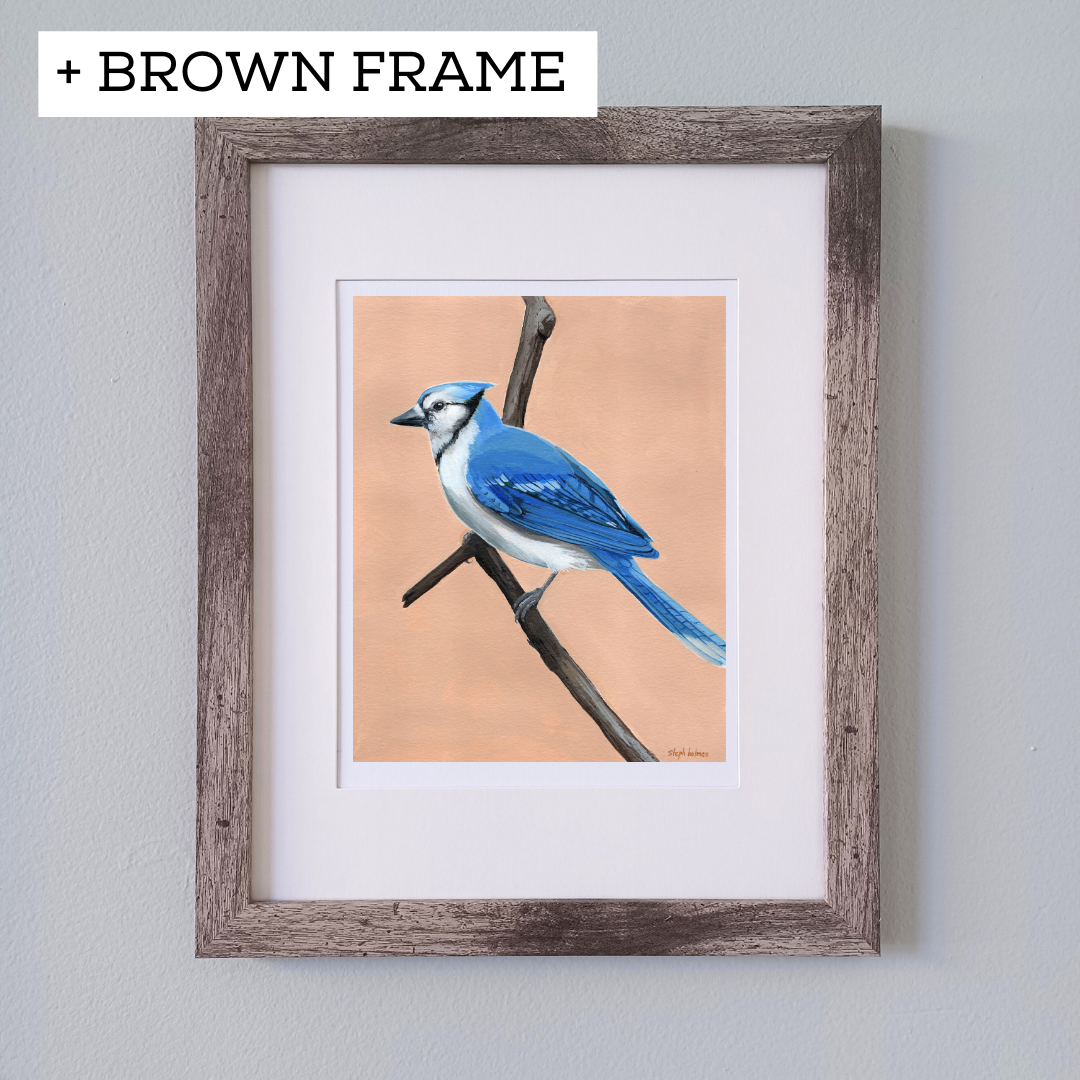 An art print featuring a bright Blue Jay bird in a brown frame in front of a white wall.