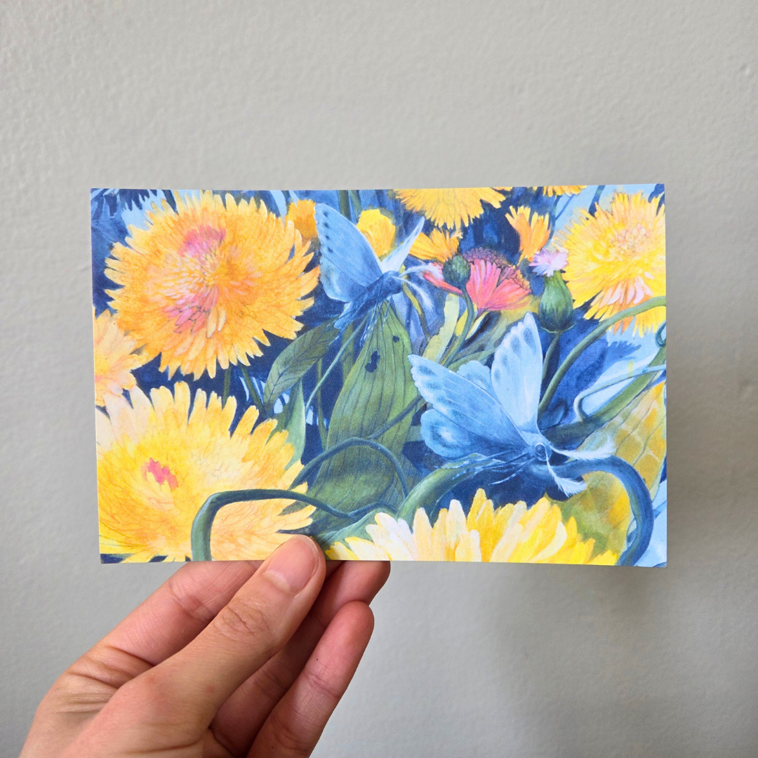 A hand holding an art print postcard featuring moths and dandelions.