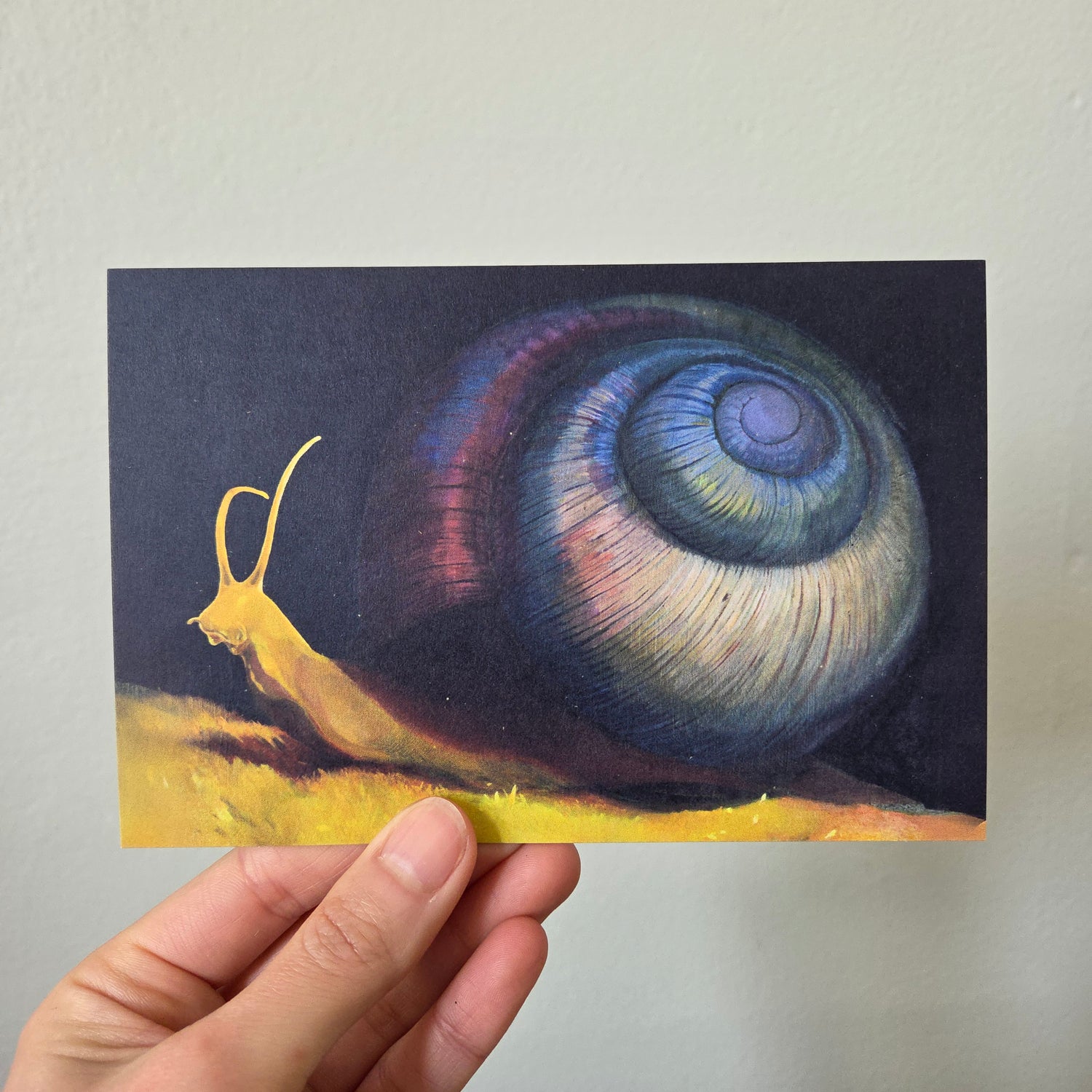 A hand holding an art print postcard featuring a purple snail.